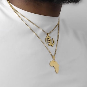 May include: Two gold-toned necklaces worn together. The shorter chain features a gold-toned Adinkra symbol pendant, while the longer chain displays a gold-toned pendant shaped like the continent of Africa. Both necklaces have fine link chains.