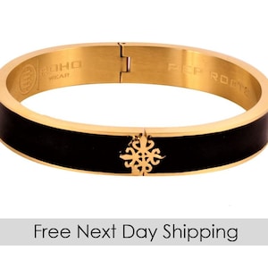 Puede incluir: Pulsera de brazalete de oro con una banda de esmalte negro y un diseño decorativo dorado. La pulsera está grabada con "ROHO WEAR" y "ACP ROOTS".