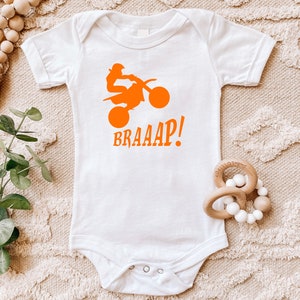 May include: White baby bodysuit with an orange graphic of a motocross rider on a motorcycle. The word "BRAAAP!" is printed below the graphic in orange. The bodysuit has short sleeves and a snap closure.