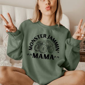 May include: Sage green sweatshirt with the text "Monster Jammin' Mama" in black, featuring a monster truck graphic. The sweatshirt has long sleeves and a crew neck. The image shows a person wearing the sweatshirt.