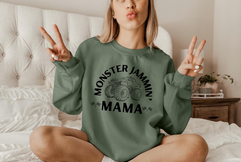 May include: Sage green sweatshirt with the text "Monster Jammin' Mama" in black, featuring a monster truck graphic. The sweatshirt has long sleeves and a crew neck. The image shows a person wearing the sweatshirt.