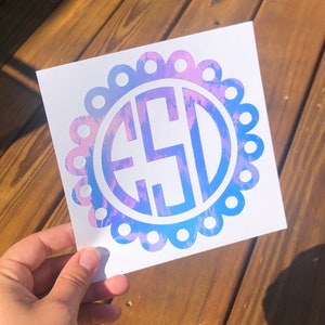 May include: A white sticker with a blue and pink iridescent monogram in a circle with a scalloped edge.
