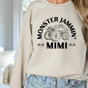 May include: Beige sweatshirt with the text "MONSTER JAMMIN', MIMI" in black, featuring a monster truck graphic. The design includes checkered flags on either side of "MIMI". The sweatshirt is a casual, comfortable style.