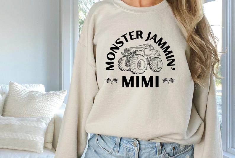 May include: Beige sweatshirt with the text "MONSTER JAMMIN', MIMI" in black, featuring a monster truck graphic. The design includes checkered flags on either side of "MIMI". The sweatshirt is a casual, comfortable style.