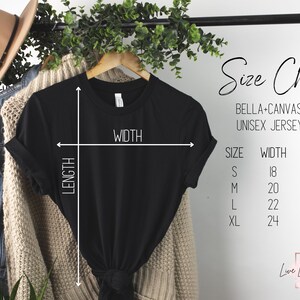 May include: A black Bella+Canvas 3001 unisex jersey tee shirt hanging on a wooden hanger. The image includes a size chart with measurements in inches: S (18x28), M (20x29), L (22x30), XL (24x31). A tan hat and denim jacket are also visible.