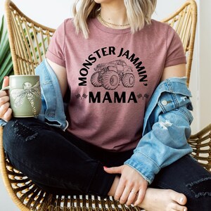 May include: A mauve t-shirt with the text "MONSTER JAMMIN', MAMA" and a monster truck graphic. The person is wearing a denim jacket and holding a green mug with a dragonfly design. The person is sitting in a wicker chair.