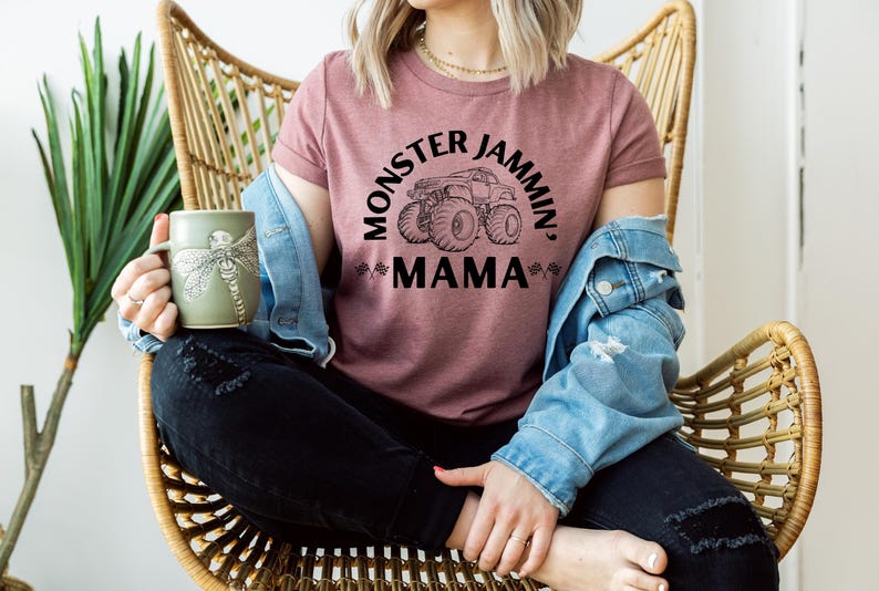 May include: A mauve t-shirt with the text "MONSTER JAMMIN', MAMA" and a monster truck graphic. The person is wearing a denim jacket and holding a green mug with a dragonfly design. The person is sitting in a wicker chair.