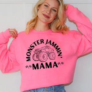 May include: A bright pink sweatshirt with the text "MONSTER JAMMIN', MAMA" in black, along with a graphic of a monster truck. The sweatshirt is a crew neck style.