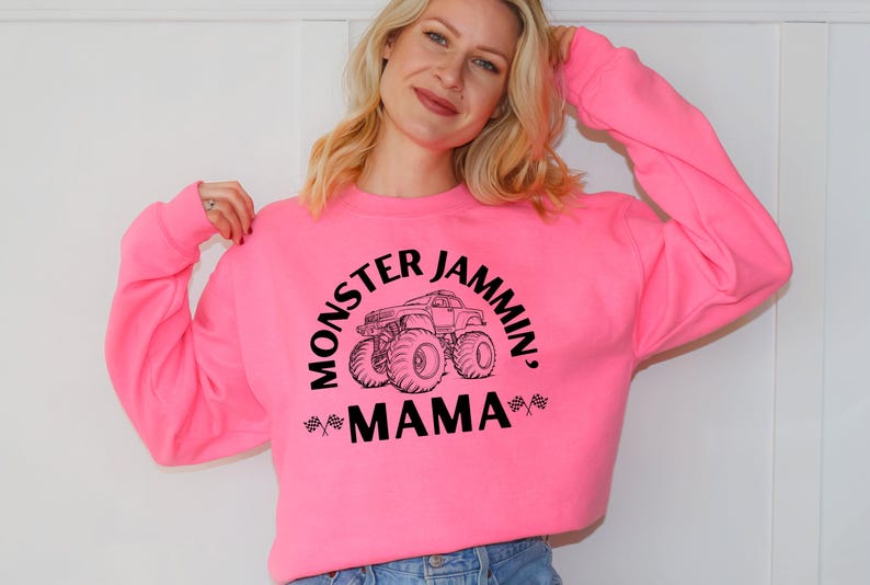 May include: A bright pink sweatshirt with the text "MONSTER JAMMIN', MAMA" in black, along with a graphic of a monster truck. The sweatshirt is a crew neck style.
