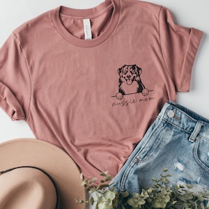 May include: A pink t-shirt featuring a black graphic of an Australian Shepherd dog peeking over the top with the text "Aussie Mom" underneath.