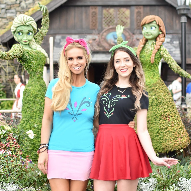 Op de afbeelding: Twee vrouwen dragen blauwe en zwarte T-shirts met een Frozen-ontwerp, roze en rode rokken en hoofdbanden. Ze staan voor twee topiary-figuren van Elsa en Anna uit Frozen.