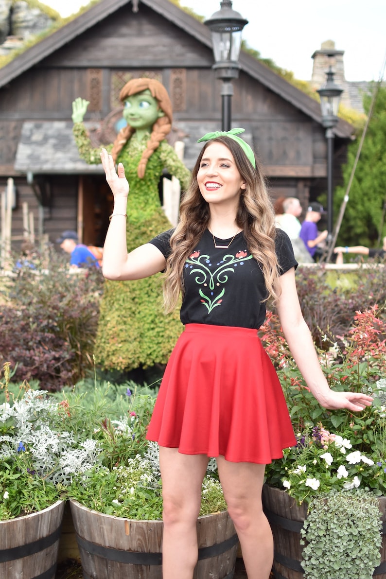 Op de afbeelding: Een vrouw in een zwart T-shirt met een groen bloemenontwerp en een rode rok staat voor een topiary-beeld van een vrouw met lang bruin haar. De vrouw glimlacht en zwaait. Het beeld staat voor een houten gebouw met een groen dak.