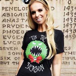 Puede incluir: Una camiseta negra con un dise&ntilde;o de corona navide&ntilde;a verde y roja que presenta un monstruo con una sonrisa de dientes. El texto "'Tis the Season" est&aacute; impreso debajo de la corona. La persona que lleva la camiseta tambi&eacute;n lleva una falda de rayas blancas y negras y una diadema con orejas de Mickey Mouse negras y una cara de Jack Skellington.