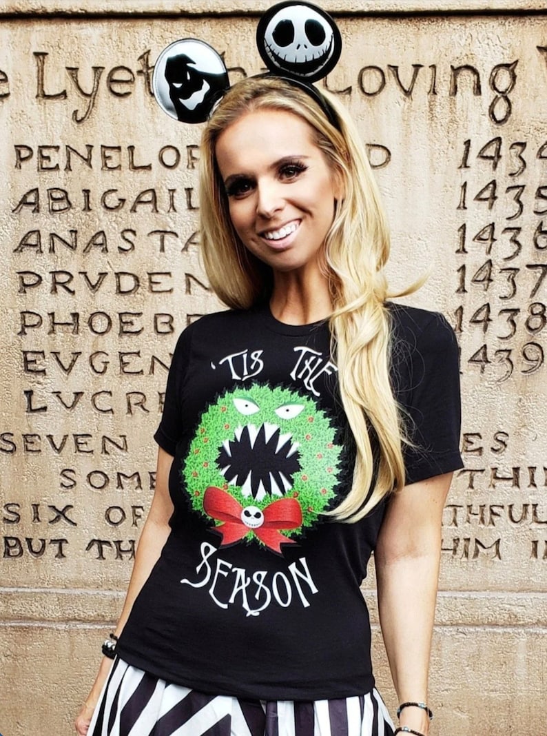 Puede incluir: Una camiseta negra con un dise&ntilde;o de corona navide&ntilde;a verde y roja que presenta un monstruo con una sonrisa de dientes. El texto "'Tis the Season" est&aacute; impreso debajo de la corona. La persona que lleva la camiseta tambi&eacute;n lleva una falda de rayas blancas y negras y una diadema con orejas de Mickey Mouse negras y una cara de Jack Skellington.