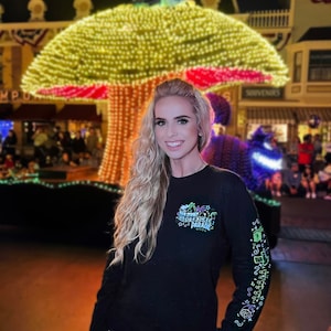 Op de afbeelding: Een vrouw draagt een zwart shirt met lange mouwen met een kleurrijke afbeelding waarop "Mickey's Very Merry Electrical Parade" staat. Ze staat voor een groot, verlicht paddenstoelvormig bouwwerk.