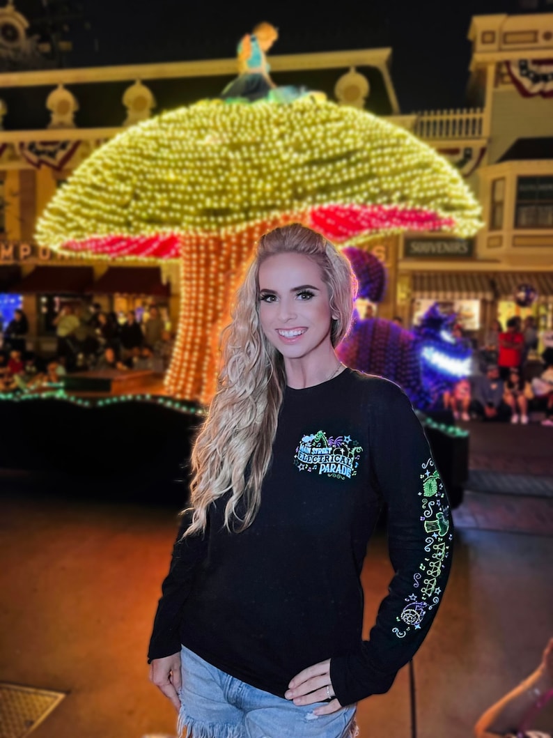 Op de afbeelding: Een vrouw draagt een zwart shirt met lange mouwen met een kleurrijke afbeelding waarop "Mickey's Very Merry Electrical Parade" staat. Ze staat voor een groot, verlicht paddenstoelvormig bouwwerk.