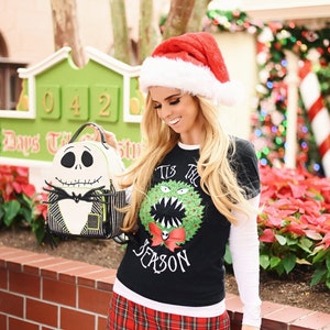 Puede incluir: Una mujer con un gorro de Pap&aacute; Noel rojo y una camisa de manga larga de color negro y blanco con una corona de Navidad verde y el texto "Tis the Season" en ella. Lleva una mochila de color negro y blanco con un dise&ntilde;o de Jack Skellington. Est&aacute; de pie frente a un cartel verde con los n&uacute;meros 042 en &eacute;l.