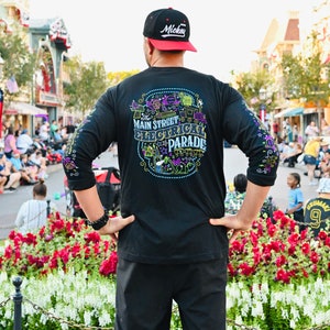 Op de afbeelding: Een persoon draagt een zwart shirt met lange mouwen met een kleurrijk grafisch ontwerp waarop "The Main Street Electrical Parade" staat. De persoon draagt een zwarte baseballpet met het woord "Mickey" in witte letters op de voorkant. De persoon staat voor een bloembed met rode en witte bloemen.