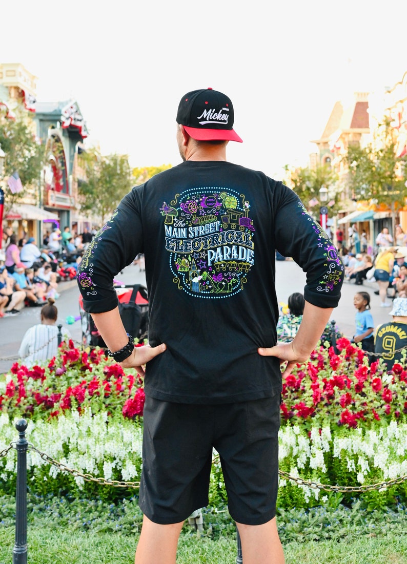 Op de afbeelding: Een persoon draagt een zwart shirt met lange mouwen met een kleurrijk grafisch ontwerp waarop "The Main Street Electrical Parade" staat. De persoon draagt een zwarte baseballpet met het woord "Mickey" in witte letters op de voorkant. De persoon staat voor een bloembed met rode en witte bloemen.