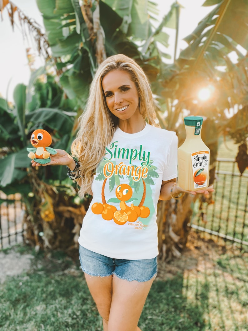 May include: A woman wearing a white t-shirt with a cartoon orange bird and the text "Simply Orange" and the words "100% Premium Florida Magic" printed on it. She is holding a plush orange bird and a bottle of Simply Orange juice.