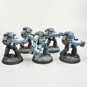May include: Five miniature figures of armoured soldiers, each holding a futuristic rifle. The figures are painted in shades of grey and blue, with detailed armour and weaponry. They stand on textured, circular bases, suggesting a tabletop game or collectible.