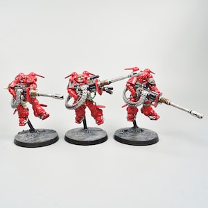 May include: Three miniature figurines of red armored soldiers, each holding a silver and black weapon. The figures stand on textured, dark gray bases. The soldiers are posed as if in action, with detailed armor and weaponry.