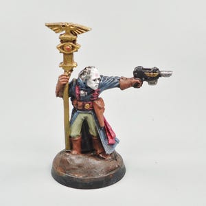 Puede incluir: Figura en miniatura de un personaje con abrigo azul, guantes marrones y pantalones verdes, sosteniendo un bastón dorado con un emblema de águila y una pistola negra. La figura está sobre una base marrón, con la cara pálida y pelo blanco.
