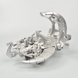 Könnte beinhalten: Ein silbernes Modell eines futuristischen Fahrzeugs mit detailliertem Design. Es verfügt über einen gebogenen Bogen und eine Plattform mit kleinen humanoiden Figuren. Das Fahrzeug hat ein schlankes, metallisches Design, das auf ein Science-Fiction- oder Fantasy-Thema hindeutet.