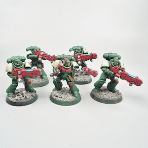 May include: Five miniature figurines of space marines in green armour with red weapons. The figurines are posed on small, textured bases. The space marines have white shoulder pads against a white background.