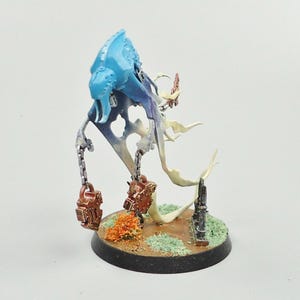May include: A miniature fantasy figure of a spectral creature with a blue head and flowing white and blue body. It has two chains with brown lockets, and stands on a base with a small bush and a tombstone.