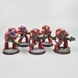 May include: Six red and black miniature figurines of armored soldiers, each holding a weapon with a neon green barrel. The soldiers are posed on grey circular bases. The helmets are blue and red.