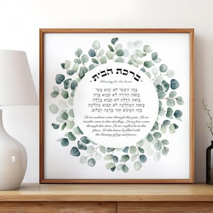 May include: A framed print with a wooden frame features a circular eucalyptus wreath surrounding text in Hebrew and English. The text is a blessing for the home, with a white background.