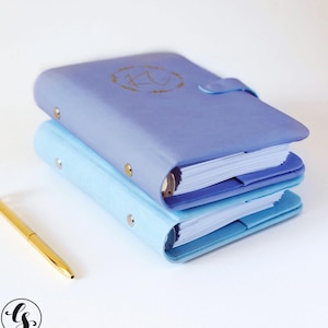 May include: Two blue leather-bound journals with gold accents and a gold initial on the top journal. The journals are stacked on top of each other.