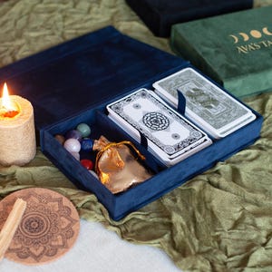 Peut inclure: Un coffret de tarot en velours bleu marine ouvert, révélant un jeu de cartes, une petite pochette dorée et des pierres précieuses colorées. Une bougie allumée et un dessous de verre en liège avec un motif mandala sont à proximité.
