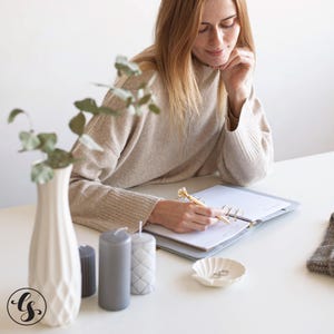 May include: A beige sweater, a white textured vase with eucalyptus, three cylindrical candles in gray shades, a gray ring-bound planner, a gold pen with a diamond topper, and a small white shell-shaped dish containing two silver rings.