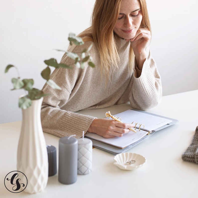 May include: A beige sweater, a white textured vase with eucalyptus, three cylindrical candles in gray shades, a gray ring-bound planner, a gold pen with a diamond topper, and a small white shell-shaped dish containing two silver rings.