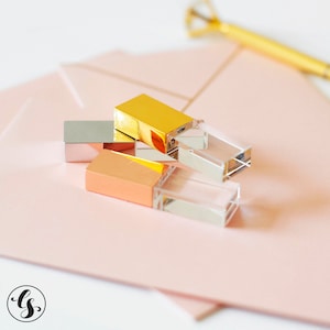 May include: Three crystal USB flash drives with gold and rose gold metal accents. The drives are stacked on top of each other on a pink surface.