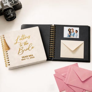 Peut inclure: Un album à spirales avec une couverture crème portant le texte doré Letters to the Bride, DEAR MIA, MAY 17, 2026, et un pompon doré. Les pages noires ouvertes présentent une photographie de trois personnes et une enveloppe crème.