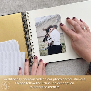 May include: An open scrapbook with a gold-colored spine and pages. A photograph of a couple is placed on a page. The photo is held in place by a person's hand with red nail polish. Additional white photo corners are visible.