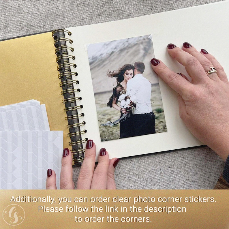 May include: An open scrapbook with a gold-colored spine and pages. A photograph of a couple is placed on a page. The photo is held in place by a person's hand with red nail polish. Additional white photo corners are visible.