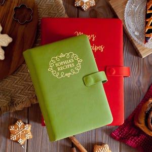 May include: A green recipe book with a snap closure and the text "SOPHIA'S RECIPES" in gold, layered over a red recipe book with a snap closure and gold text. Both books feature a smooth, matte finish and are rectangular in shape.