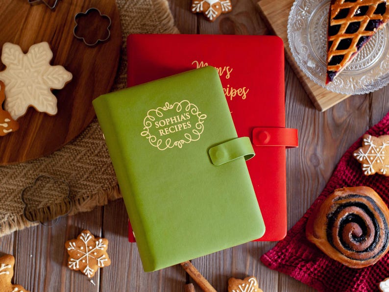 May include: A green recipe book with a snap closure and the text "SOPHIA'S RECIPES" in gold, layered over a red recipe book with a snap closure and gold text. Both books feature a smooth, matte finish and are rectangular in shape.