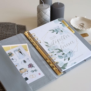 May include: A gray wedding planner with gold accents, open to a page with the text "Our Together is Forever." The planner includes stickers and tabs labeled "Wedding Day" and "Inspiration."