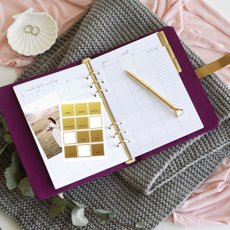 May include: A deep purple ring-bound planner with white monthly calendar pages, a gold-toned pen with a diamond-shaped topper, a sheet of gold-colored adhesive labels, and a photograph of a person in a white dress standing in a field.