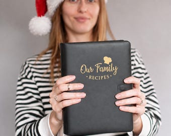 May include: A black rectangular recipe binder with a gold-colored chef hat icon and the text Our Family RECIPES printed on the front cover. The binder features a smooth matte finish and a small black tab closure on the right side edge.