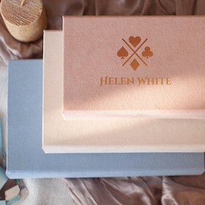May include: Three stacked, rectangular boxes in pastel colors: light blue, cream, and pink. The top box is pink with a gold logo and the name "HELEN WHITE". A candle and tarot cards are in the background. The boxes appear to be for storage or presentation.