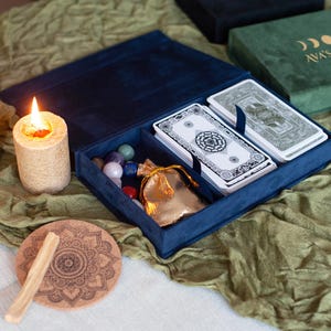 May include: A blue velvet tarot card box with a deck of cards, a small pouch, and crystals inside. The box is open and the cards are visible. The box is sitting on a green fabric surface.