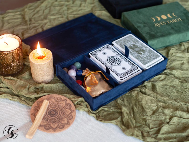 May include: A blue velvet tarot card box with a deck of cards, a small pouch, and crystals inside. The box is open and the cards are visible. The box is sitting on a green fabric surface.