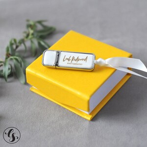 K&ouml;nnte beinhalten: Ein wei&szlig;er USB-Stick mit silbernem Metallgeh&auml;use und einer goldenen Aufschrift "Leah Redmond Photography" in handschriftlicher Schrift. Der Stick liegt auf einer gelben Box mit einem wei&szlig;en Band.