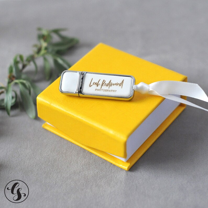 K&ouml;nnte beinhalten: Ein wei&szlig;er USB-Stick mit silbernem Metallgeh&auml;use und einer goldenen Aufschrift "Leah Redmond Photography" in handschriftlicher Schrift. Der Stick liegt auf einer gelben Box mit einem wei&szlig;en Band.